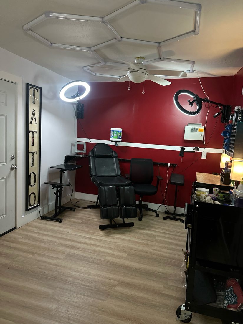 The tattoo side of the studio: a black tattoo chair under a vertical TATTOO sign and ring light, same crimson wall
